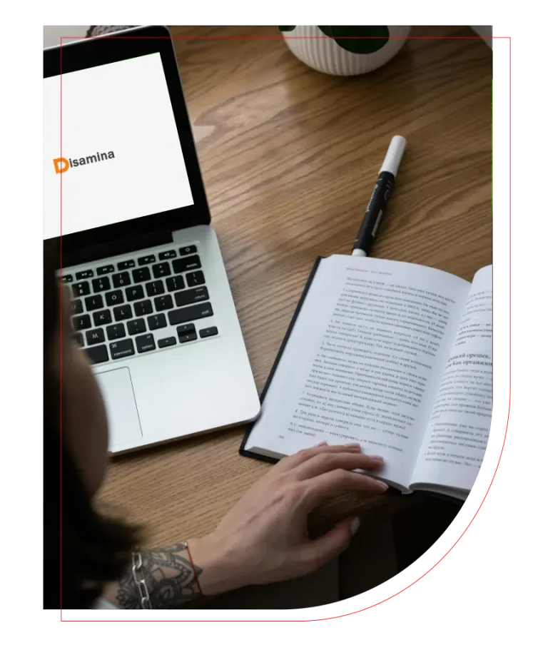 Person reading a book beside an open laptop on a wooden table.